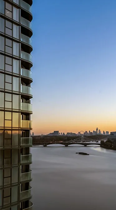 Dawn breaks over London, with the silhouette of a modern skyscraper on the left and the serene River Thames flowing towards a distant city skyline, featuring the silhouettes of iconic buildings under a clear gradient sky.