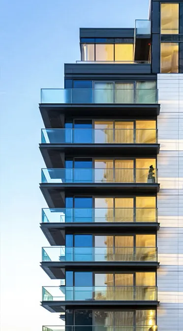 The evening sun reflects off the glass balconies of a modern apartment building, showcasing the clean lines and reflective surfaces of its architecture, with a clear blue sky in the background creating a contrast that highlights the building’s urban design.