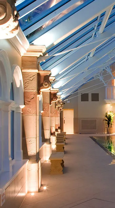 Illuminated poolside corridor with arched windows and ornate pillars under a glass roof.