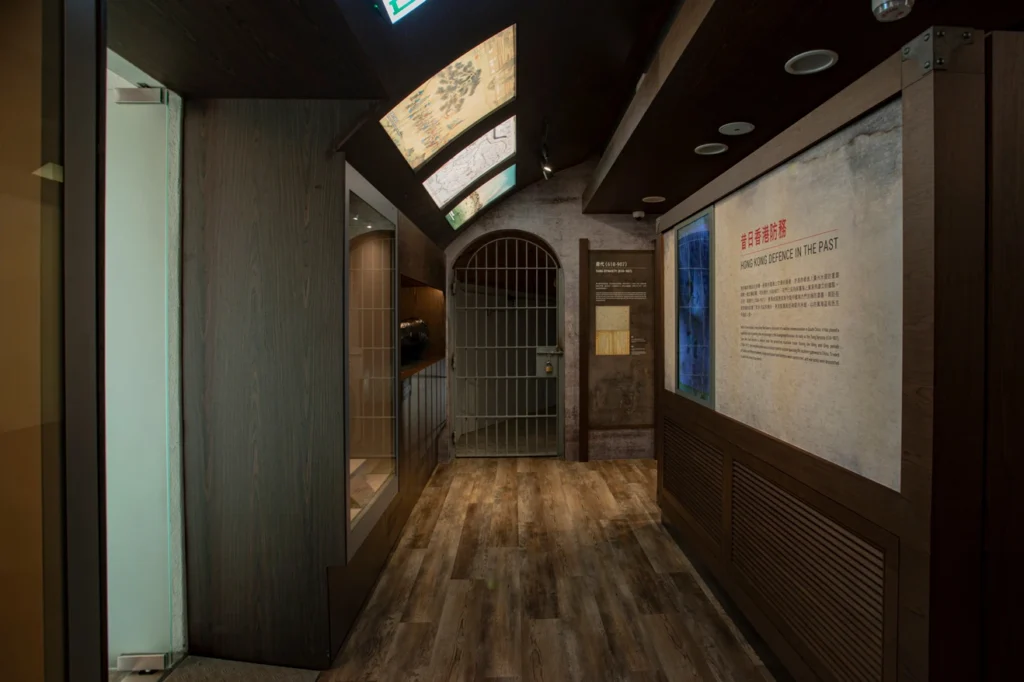 Dimly lit gallery with wooden flooring, interpretive panels, and a pirate jail gate depicting Hong Kong's defence history at the Museum of Coastal Defence