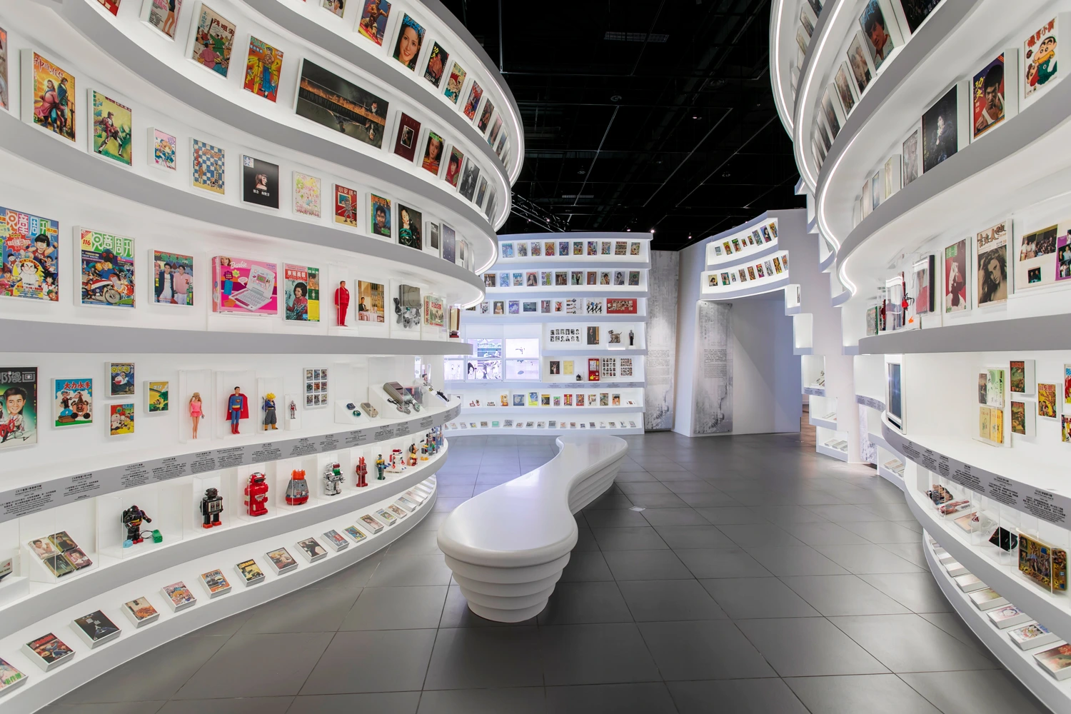 A sweeping, white display wraps around the room with ascending shelves filled with colorful memorabilia, comic books, toys, and collectibles, creating an immersive timeline of Hong Kong’s entertainment history.