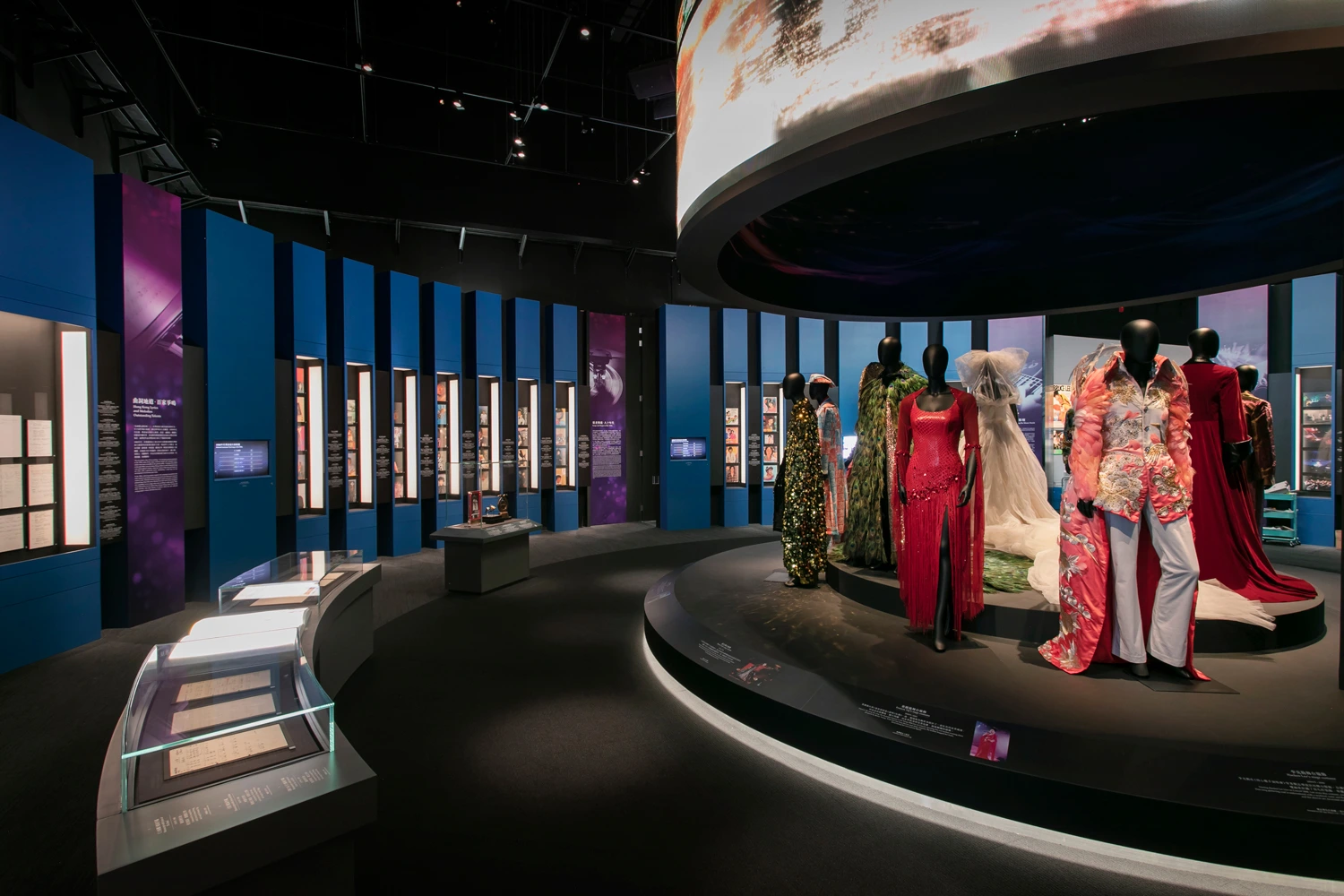 The exhibit features a central circular stage showcasing a collection of exquisite costumes, surrounded by blue vertical panels with informational content, highlighting Rebel Arkitekt's project management in bringing to life the theatrical heritage as part of Hong Kong's vibrant cultural scene.